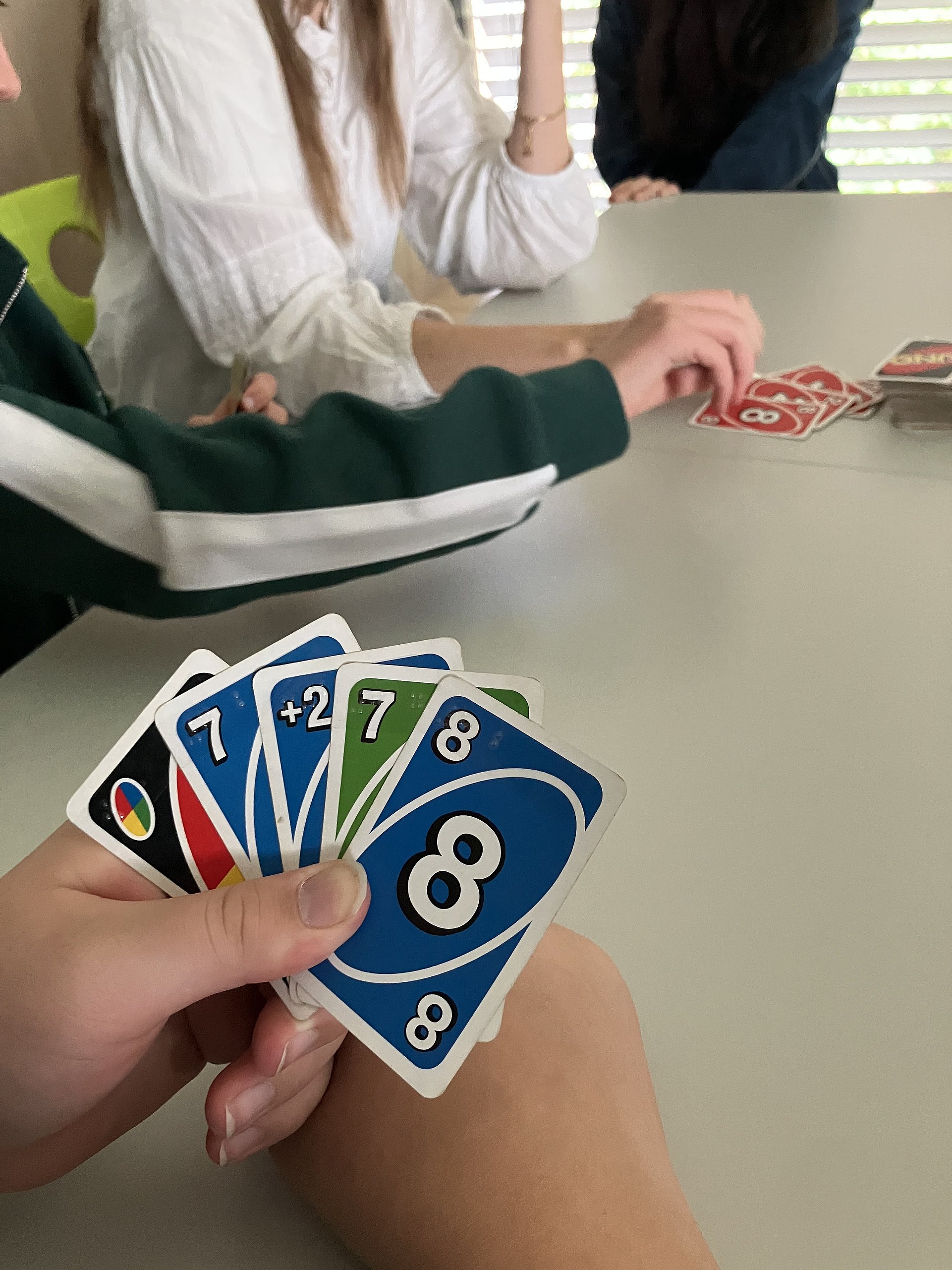 Eine Hand hält ein Satz bunter Uno-Karten. An einem Tisch sitzen Menschen zusammen und spielen Karten