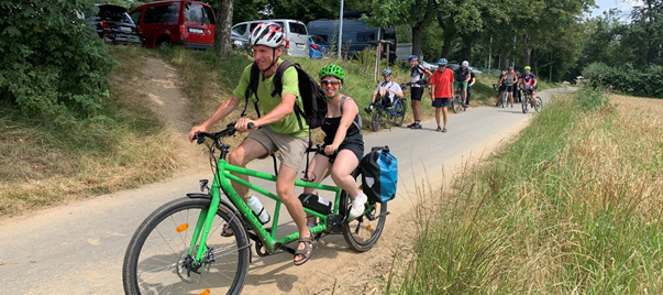 Zwei Menschen fahren zusammen auf einem grünen Tandem eine Straße entlang. Sie lächeln in die Kamera.