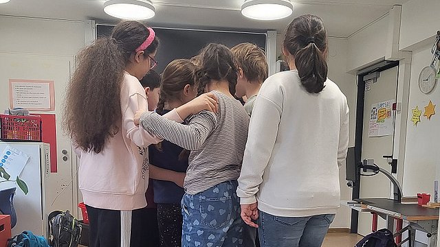 Mehrere Kinder stehen gemeinsam auf einer kleinen Holzbrücke im Klassenzimmer. Sie halten sich aneinander fest mit dem Rücken zur Kamera.