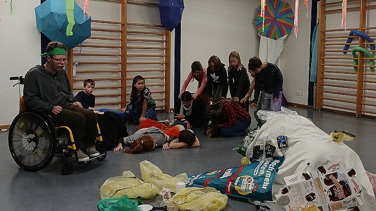 Mehrere Kinder und Jugendliche in schwarzer Kleidung und ein Junge im Rollstuhl stehen nebeneinander in einem Raum. Einige von ihnen sind auf den Knien und nach vorne gebeugt. Vor ihnen liegt in der Mitte ein Berg aus Plastikmüll.