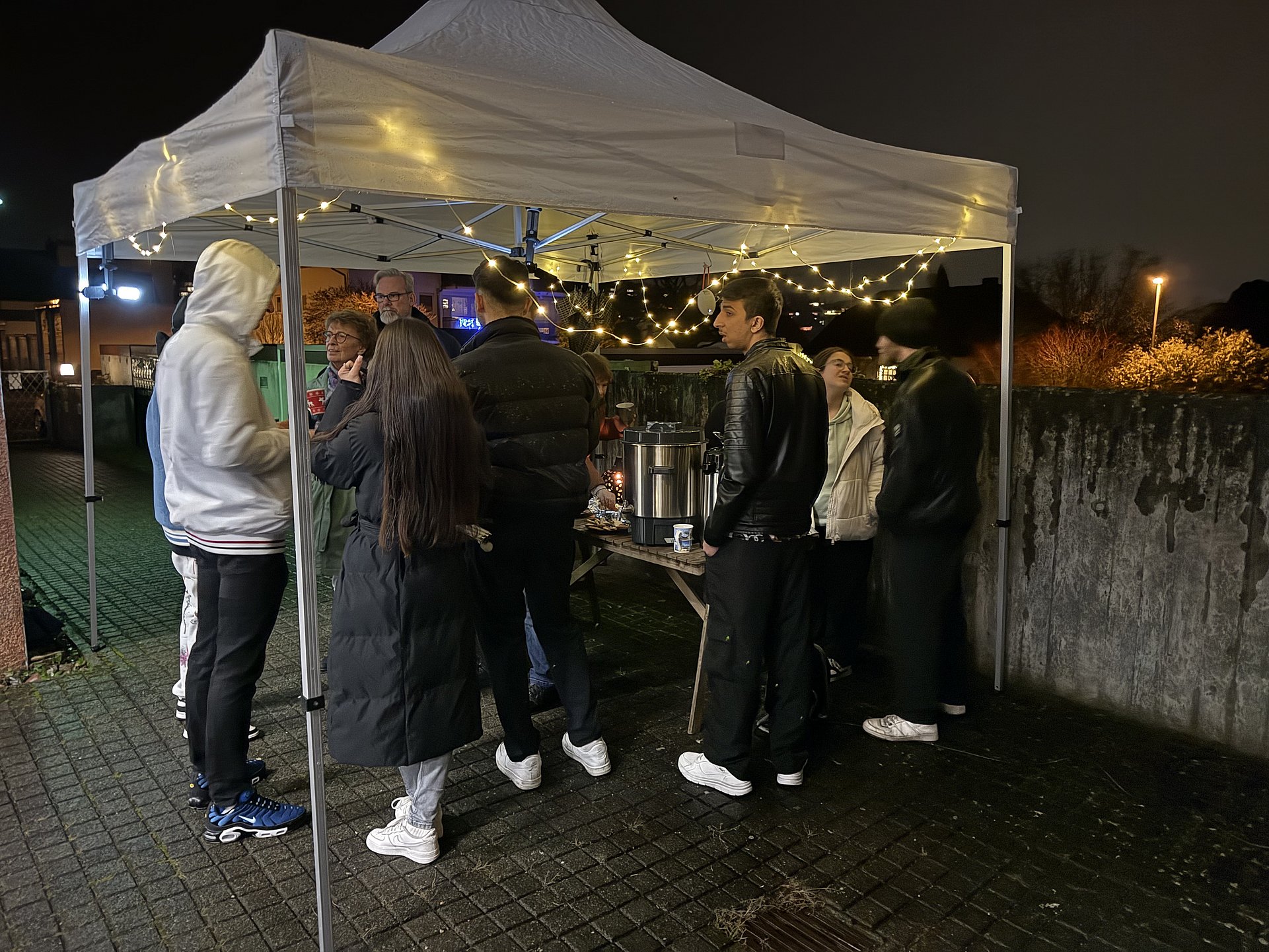 Auf einem Gehweg steht ein offenes weißes Zelt. Darunter stehen Personen nebeneinander und unterhalten sich. Das Zelt ist mit einer Lichterkette geschmückt. 