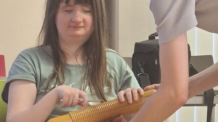 Eine junge Frau mit langen, braunen Haaren sitzt auf einem Stuhl und hält ein längliches, geriffeltes gelbes Musikinstrument in den Händen, das an eine große Holzraspel erinnert. Sie trägt ein graugrünes T Shirt und eine schwarze Hose mit hellem Muster. Ihre Hände liegen auf dem Instrument, die Finger sind so positioniert, als würde sie darüber streichen oder es spielen. Vor ihr beugt sich eine weitere Frau mit kurzen, dunklen Haaren von rechts ins Bild und reicht das Instrument mit beiden Armen. Sie trägt ein kurzärmeliges, hellgraues Oberteil. Im Hintergrund stehen ein Tisch und eine dunkle Tasche, außerdem helle Vorhänge.