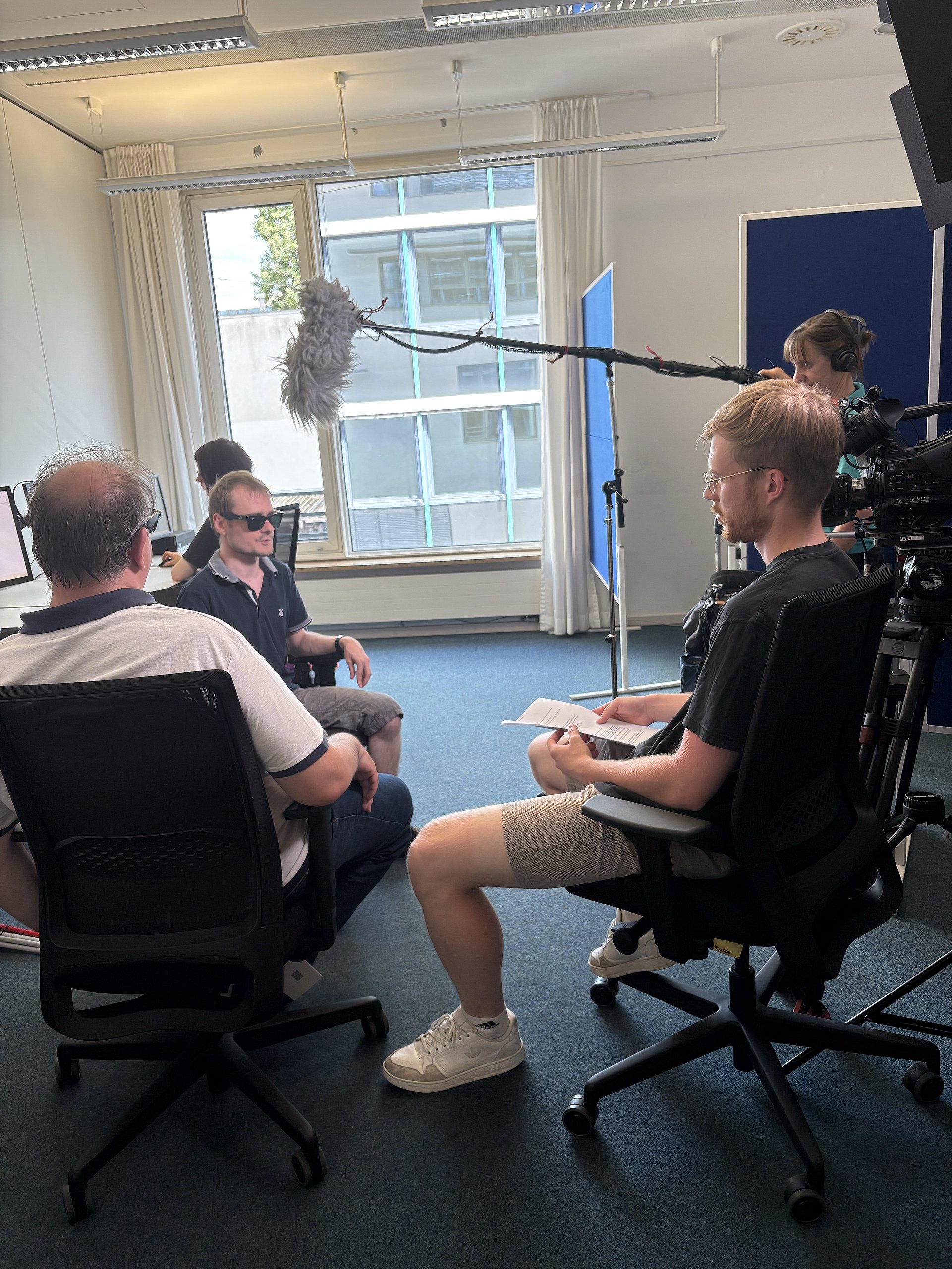 Fünf Personen in einem Unterrichtsraum. Links ein Mann in Jeans und weißem T-Shirt, der auf einem Schreibtischstuhl sitzt und nach vorne weg von der Kamera blickt. Rechts hinter ihm sitzt ein junger Mann mit blauem T-Shirt und schwarzer Brille in einem Stuhl. Rechts vorne sitzt ein Mann in kurzer Hose und schwarzem T-Shirt auf einem Stuhl, er hält einen Zettel in der Hand und blickt in Richtung des jungen Mannes. Hinter ihm steht eine Person, die ein Mikrofon an einem langen Stab über die Männer hält.