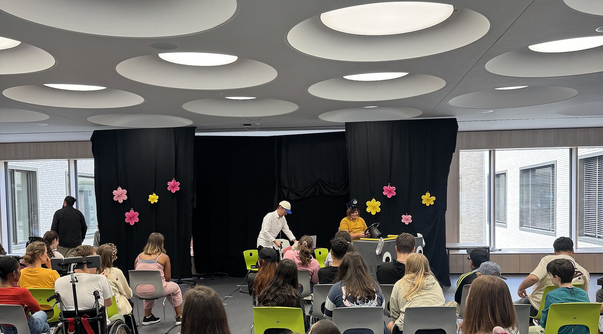 In der Aula des Schulgebäudes sitzen viele Schülerinnen und Schüler, darunter auch einige im Rollstuhl. Ganz Vorne ist eine Art Bühne zu erkennen auf der verkleidete Schüler ein Theaterstück aufführen