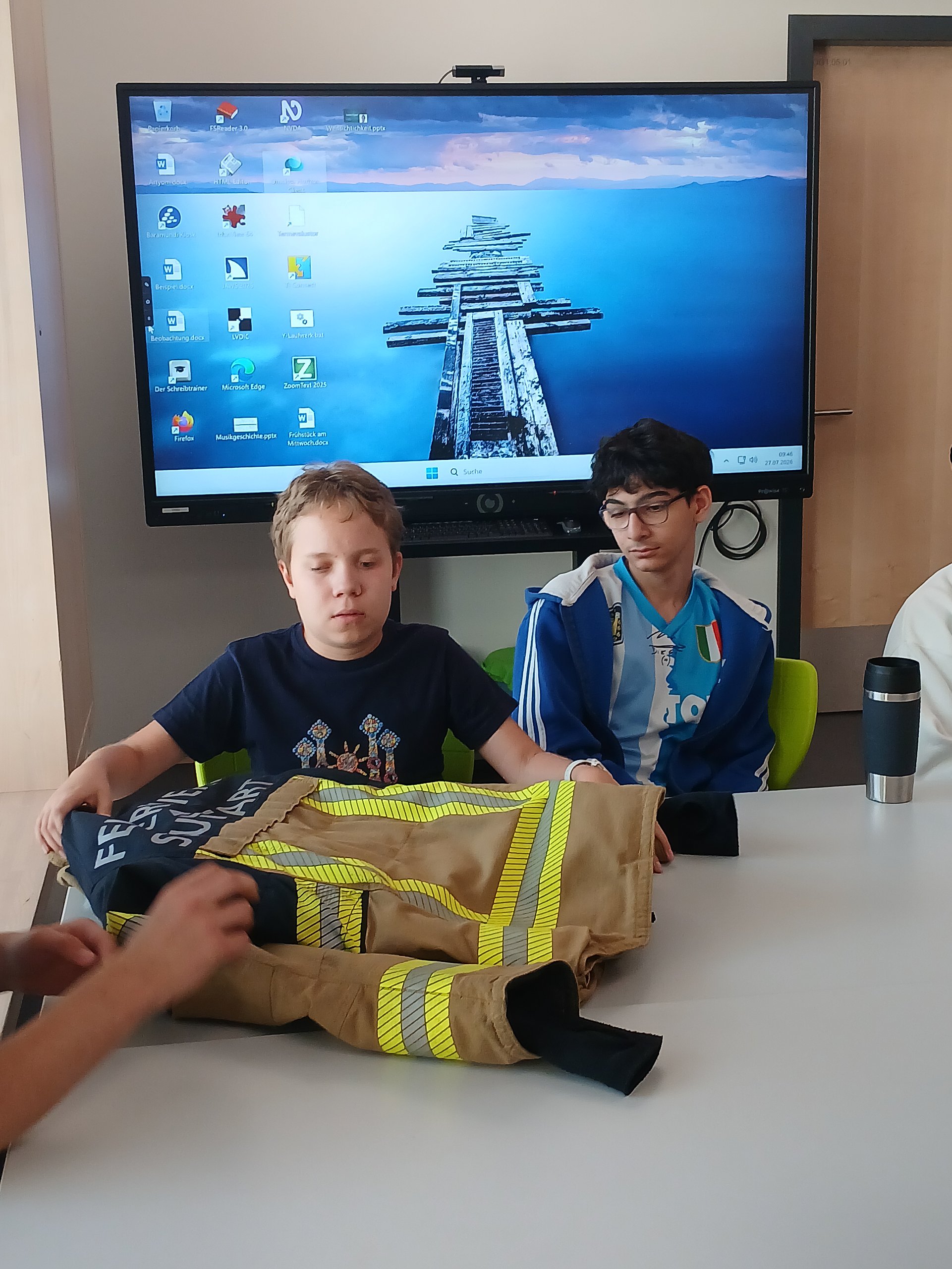 Zwei Schüler sitzen im Klassenzimmer. Ein Schüler ertastet einen Schutzanzug der Feuerwehr