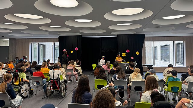 In der Aula des Schulgebäudes sitzen viele Schülerinnen und Schüler, darunter auch einige im Rollstuhl. Ganz Vorne ist eine Art Bühne zu erkennen auf der verkleidete Schüler ein Theaterstück aufführen