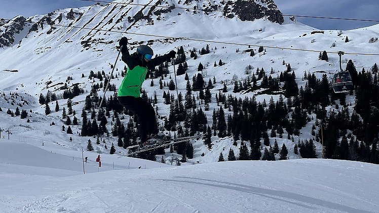 Eine Person mit Ski, Helm und grüner Skijacke fliegt in der Luft, unter ihr ist eine Skipiste.