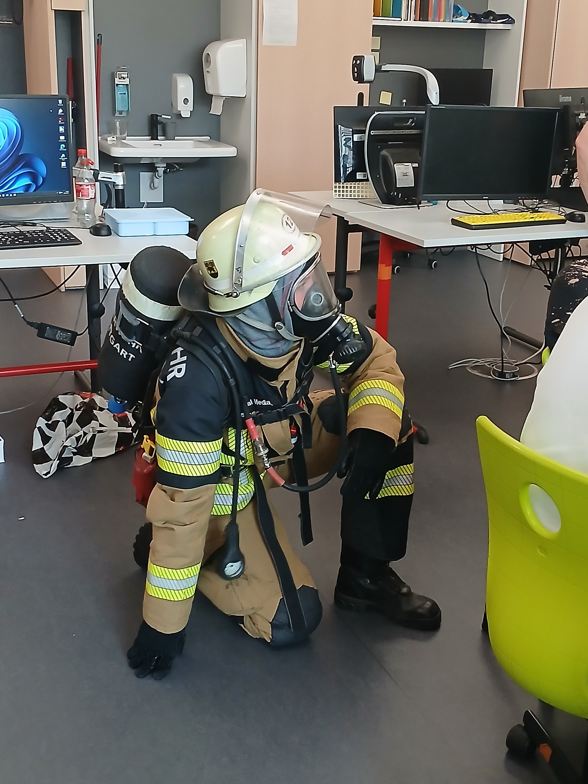 Ein Feuerwehrmann in Schutzanzug, Helm und Maske kniet auf dem Boden des Klassenzimmers