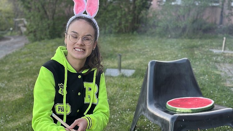 Eine junge Frau sitzt auf einem Hocker und hält einen Stock mit Stockbrot über ein Lagerfeuer vor ihr. Sie trägt einen Haarreif mit Hasenohren und einen schwarz-gelben Pullover.