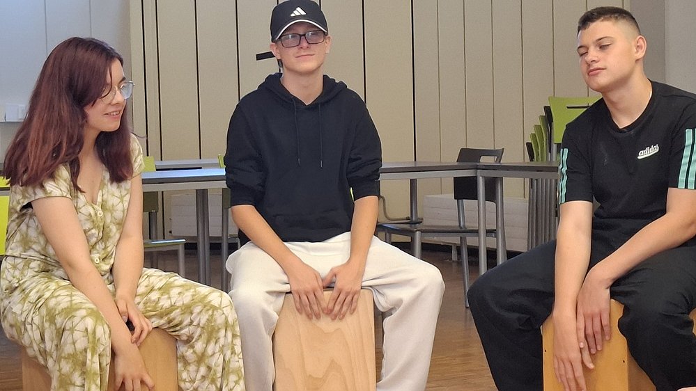 Eine junge Frau und zwei junge Männer sitzen nebeneinander auf Holzcajons in einem Innenraum. Links sitzt die junge Frau, sie hat schulterlange braun-rötliche Haare und Brille. Sie trägt eine Kombination aus Kurzarmoberteil und weiter Hose im gleichen beige-grün gemusterten Stoff. Sie sitzt leicht nach vorn gebeugt, die Unterarme ruhen locker zwischen den Knien auf der Cajon, die Hände hängen entspannt nach unten. Rechts sitzt ein junger Mann mit schwarzem Kapuzenpulli und weiter, heller Jogginghose. Er trägt eine schwarze Baseballkappe mit einem weißen Logo und eine Brille. Er sitzt aufrecht, beide Hände liegen mit gespreizten Fingern flach vorne auf der Cajon. Rechts daneben sitzt ein weiterer junger Mann, er hat kurze dunkle Haare und trägt eine schwarze Jogginghose und Pullover. Er schlägt mit beiden Händen auf die Vorderseite seines Cajons. Alle drei Cajons sind aus hellem Holz. Auf der vorderen Fläche der rechten Cajon ist unten ein kleines, dunkles Muster mit stilisierten, tanzenden Figuren zu sehen, die linke Cajon wirkt glatt ohne sichtbare Verzierung. Im Hintergrund stehen Tische und Bänke in einem Raum mit hellen Wandpaneelen.