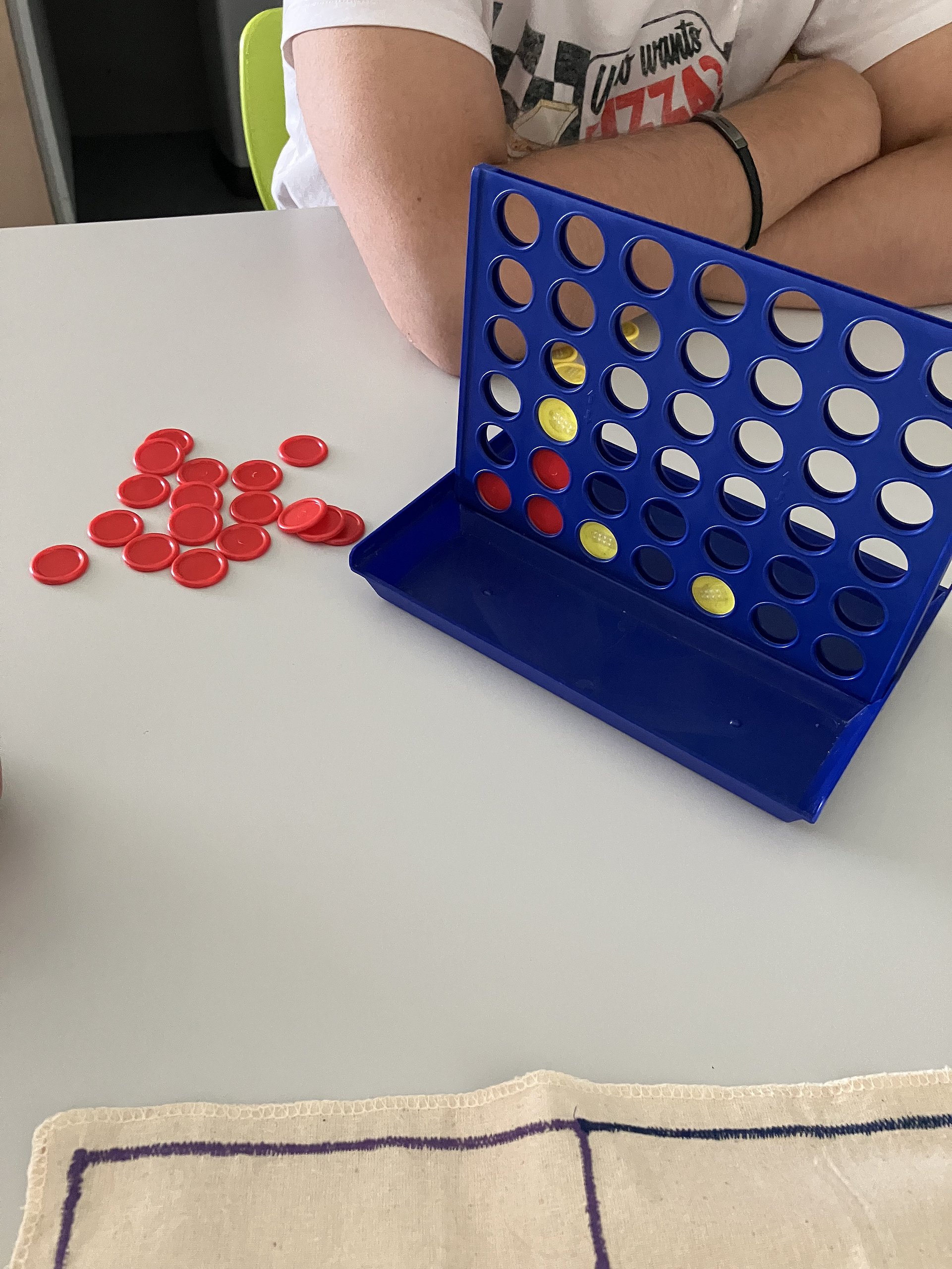 Auf einem Tisch steht ein blaues Spiel "Vier in einer Reihe". Dahinter sieht man die Arme eines Kindes