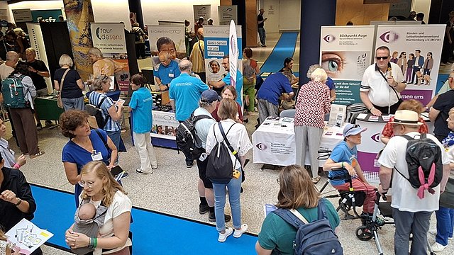 Großer Veranstaltungssaal mit vielen Menschen auf einer Messe: Mehrere Stände stehen dicht nebeneinander auf einem hellen Holzfußboden. Vorne links befindet sich ein Metallständer mit mehreren Reihen Flyern. Dahinter steht ein gelber, mit Stoff bedeckter Tisch mit Infomaterial. Darüber hängt ein großes gelbes Banner mit dem Schriftzug „Reinecker“ und dem Foto eines älteren Mannes und einer älteren Frau mit Brille, beide lächeln.  Viele Personen stehen oder gehen durch die Gänge, einige mit Rollatoren, andere im Gespräch an den Ständen. Die Menschen tragen überwiegend Freizeitkleidung wie T Shirts, kurze Hosen oder Sommerkleider. Der Raum ist hoch und modern mit grauen Wänden und einer geschwungenen Decke mit vielen kleinen Einbauleuchten. Rechts oben ist eine Empore zu erkennen. An der Decke hängen zahlreiche Scheinwerfer und technische Elemente.