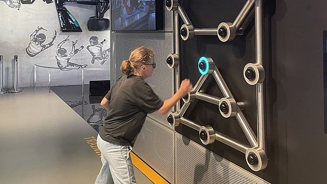 Eine Mädchen mit einem blonden Pferdeschwanz und einer Brille steht in einem Museum vor einer großen dunklen Wand. And der Wand sind runde metallische Objekte befestigt. Eines davon leuchtet blau auf. Die Objekte sind untereinander mit metallischen Leisten verbunden. Das Mädchen drückt mit ihrer rechten Hand einer der Objekte