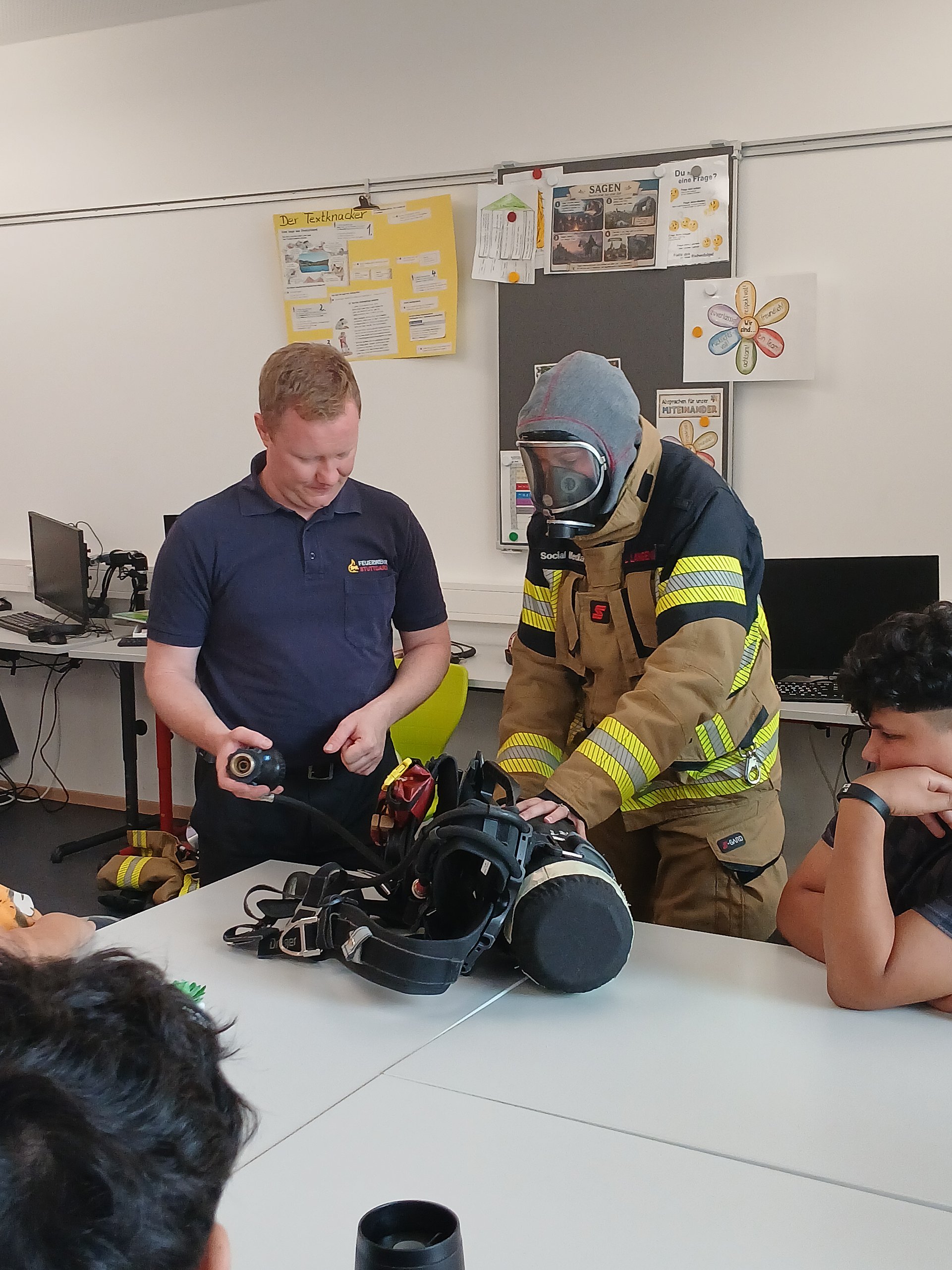 Zwei Feuerwehrleute stehen im Klassenraum und zeigen den Schülern die Ausrüstung. Eine von ihnen trägt ein Schutzanzug mit einer Gasmaske