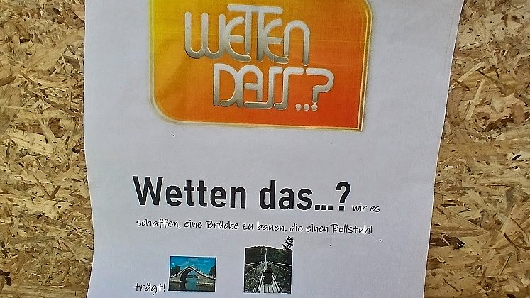 Ein Blatt Papier klebt auf einem Holzbrett. Auf dem Papier steht "Wetten dass...? wir es schaffen, eine Brücke zu bauen, die einen Rollstuhl trägt?  Wir würden uns freuen, wenn ihr zu unserer Challenger annehmt. Gerne würden wir euch zu einem kleinen Fest einladen am letzten Schultag vor den Ferien.“