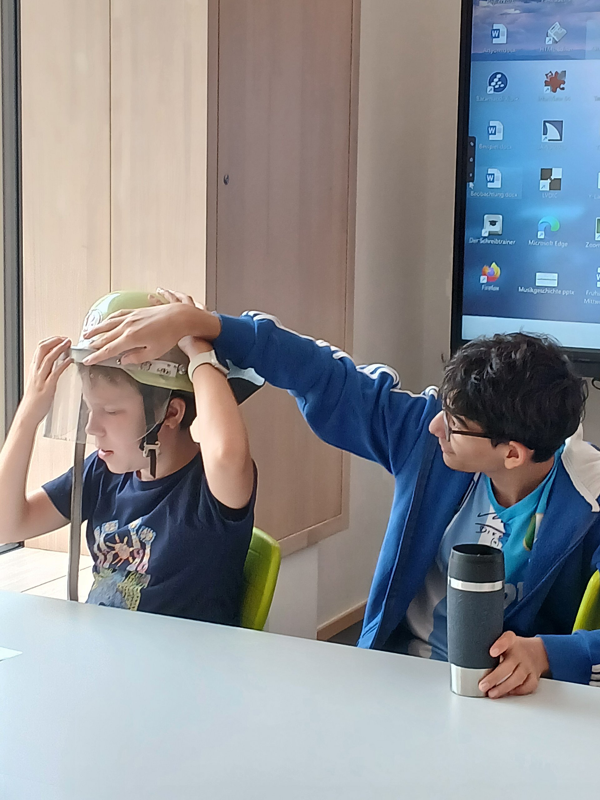 Zwei Schüler sitzen nebeneinander an einem Tisch. Der rechte trägt einen Feuerwehrhelm, der Linke betastet ihn mit der Hand