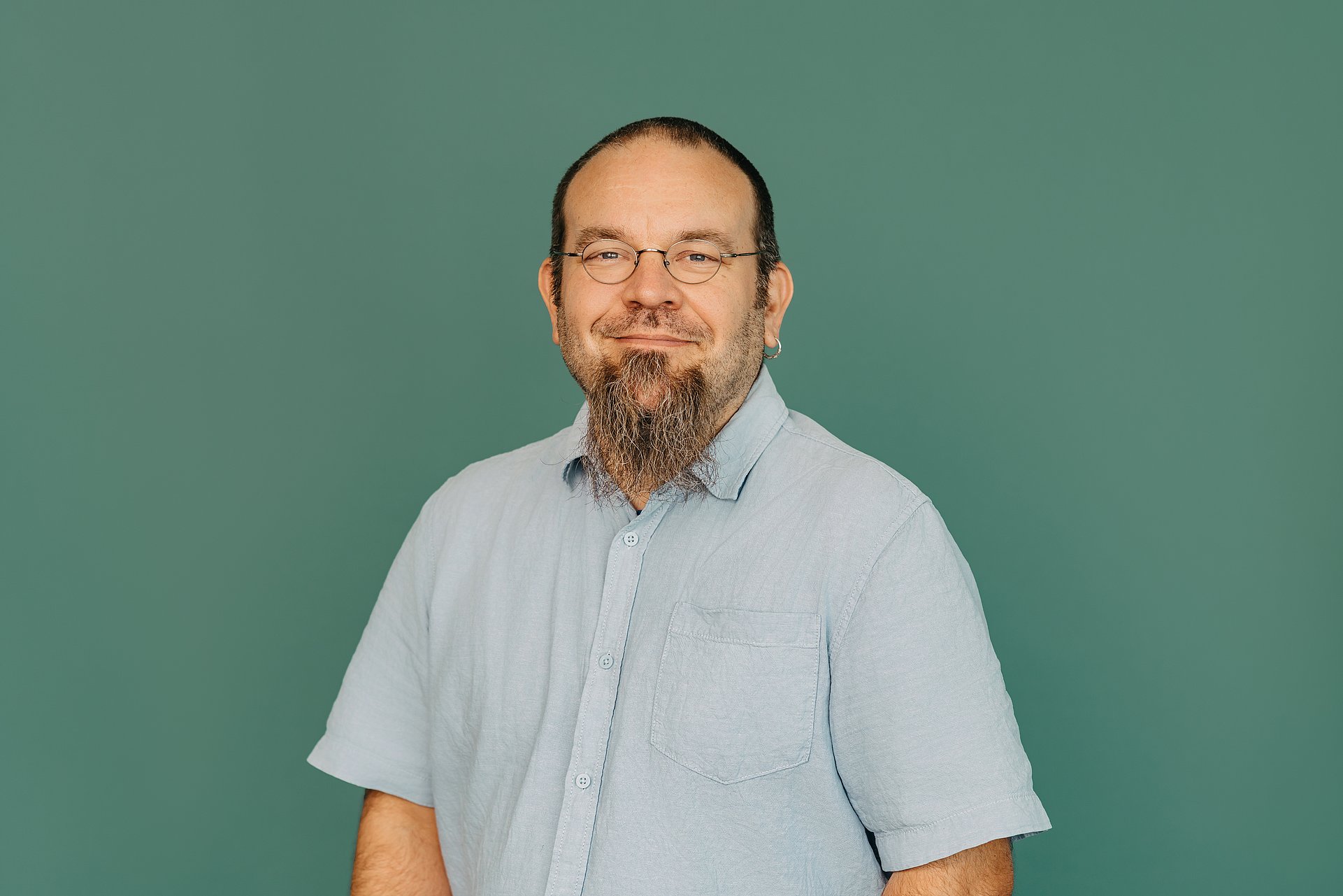 Ein Mann steht vor einem grünem Hintergrund. Er trägt ein hellblaues Hemd mit kurzen Ärmeln. Er hat kurze braune Haare und einen langen Kinnbart. Er trägt eine Brille und lächelt freundlich