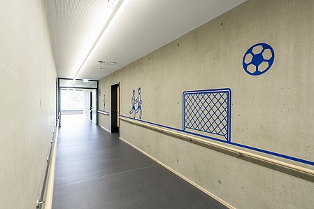 Ein Flur im barrierefreien Schulhaus des Betty-Hirsch-Schulzentrums mit Handlauf, Kontrasten, Wandtatoo zur Orientierung und länglichen Deckenleuchten