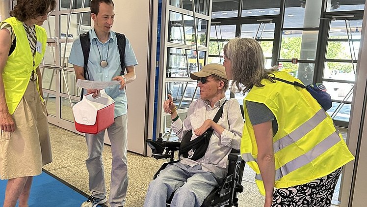 Vier Personen in einem Eingangsbereich auf einem blauen Teppich.   In der Bildmitte sitzt eine Person mit heller Haut in einem elektrischen Rollstuhl, trägt eine hellgraue Hose, helle Turnschuhe, ein helles Oberteil und eine beigefarbene Kappe. Vor der Person hängt eine schwarze Umhängetasche, in der rechten Hand hält sie ein Blatt Papier.   Links davon steht eine Frau mit heller Haut in einem gelben Warnüberwurf über einem ärmellosen Oberteil und einem knielangen beigen Rock, an den Füßen Sandalen.   Neben ihr steht ein Mann mit heller Haut in einem hellblauen Kurzarmhemd und grauer Hose, mit Rucksackgurten über den Schultern und einem Ausweisband um den Hals; er hält eine weiße Mappe und eine rot weiße Box in den Händen.   Rechts vom Rollstuhl beugt sich eine weitere Frau mit heller Haut vor; sie trägt ebenfalls eine gelbe Warnweste, ein schwarzes T Shirt und weite schwarz weiß gemusterte Hose mit flachen Sandalen. Alle scheinen miteinander zu sprechen. Im Hintergrund befinden sich große Glas- und Metalltüren eines modernen Gebäudeeingangs mit hellem Boden.