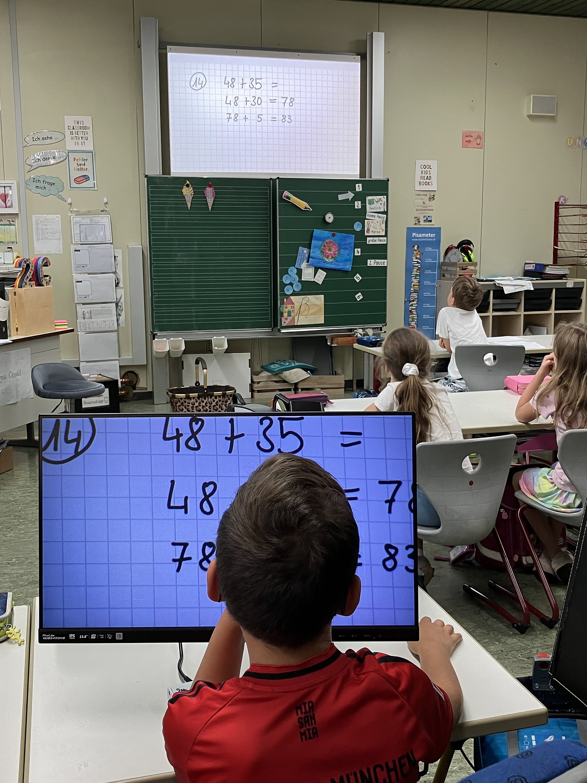 Schülerinnen und Schüler sitzen in einem Klassenzimmer und schauen nach vorne an die Tafel. Ein Schüler sitzt vor einem Bildschirm, auf dem Rechenaufgaben zu erkennen sind