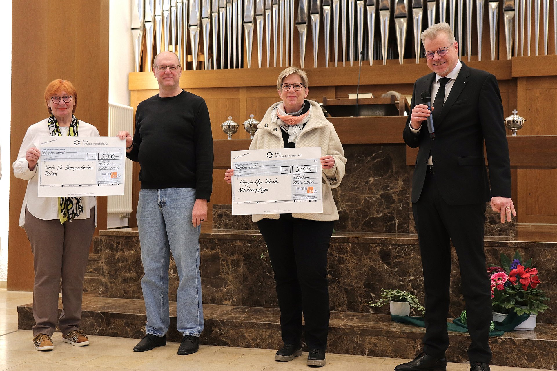 Zwei Frauen und zwei Männer stehen vor einem Altar in der Neuapostolischen Kirche. Hinter ihnen ist eine große Orgel zu sehen. Die Frauen halten einen großen Scheck in den Händen