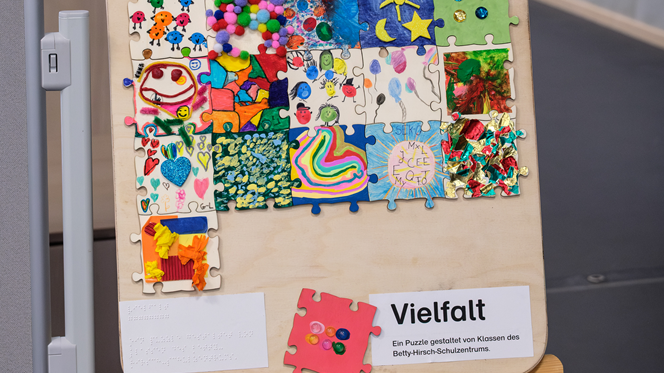 Auf einer Staffelei steht ein großes Bild, das mit Stoffen und  Farben bunt gestaltet ist. Unten rechts in der Ecke steht "Vielfalt".