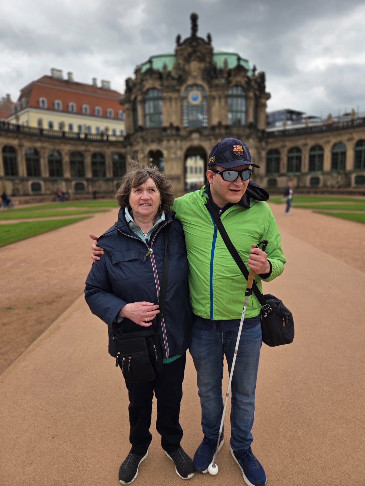 Zwei Personen stehen eng umarmt auf einem breiten sandfarbenen Weg vor einem historischen Gebäude.   Links steht eine Frau mittleren Alters mit heller Haut und mittelbraunen, leicht gewellten, kinnlangen Haaren. Sie trägt eine dunkelblaue Outdoor-Jacke mit hellblauem Futter, darunter ein helles Oberteil, eine dunkle Hose und schwarze Sportschuhe mit weißer Sohle. Über der Schulter hängt eine schwarze Umhängetasche, die seitlich vor ihrem Oberschenkel liegt. Ihr linker Arm hängt locker, mit der rechten Hand hält sie sich am Rücken der anderen Person fest und lächelt.