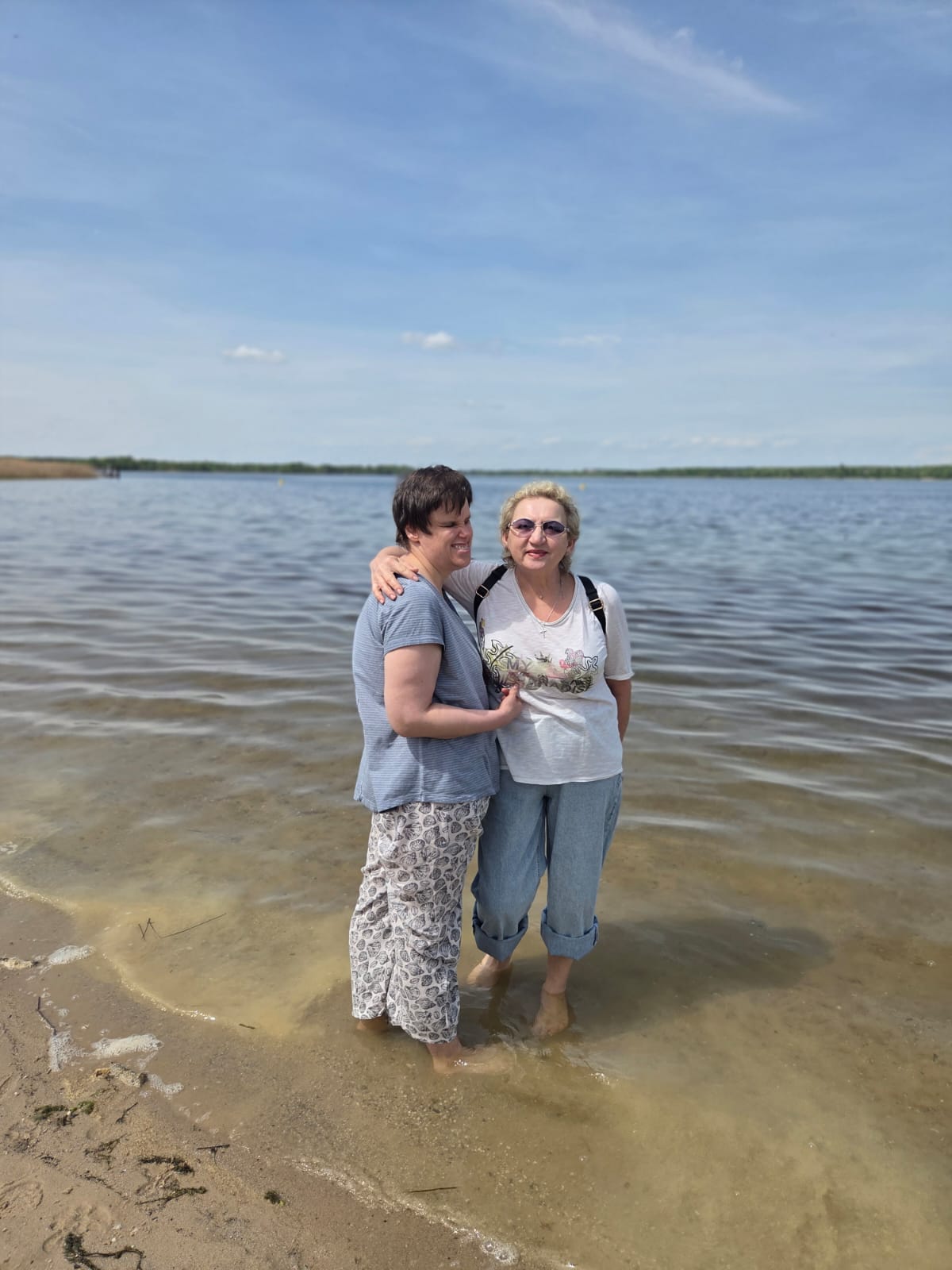 Zwei erwachsene Frauen stehen barfuß im seichten Wasser am Rand eines Sees.   Die Frau links hat kurze, dunkle Haare und eine helle Haut. Sie trägt ein hellblaues T‑Shirt und eine weiße Hose mit dunkelgemustertem Druck. Ihr Körper ist leicht zur Frau rechts gedreht, und ihr rechter Arm liegt locker um deren Rücken.  