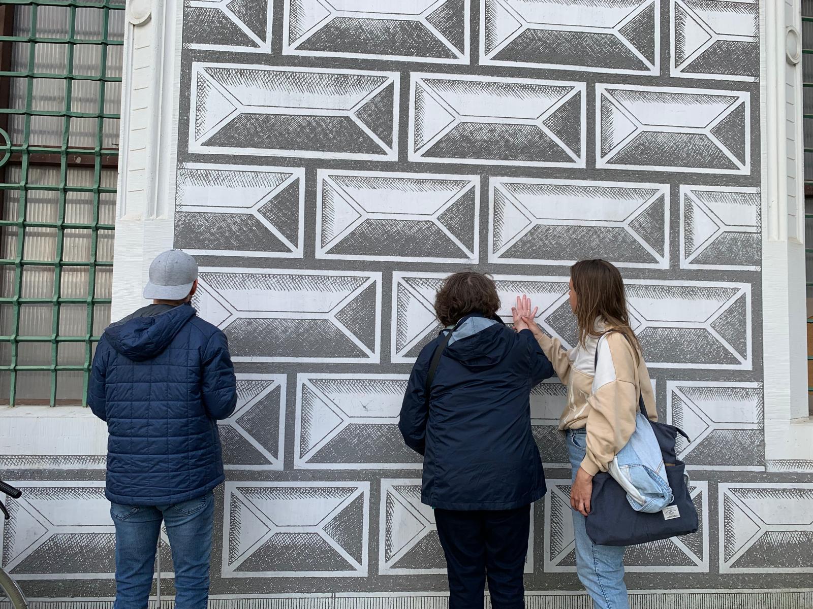 Zwei Personen stehen vor einer Wand mit grafischem Muster.   Die Wand ist mit vielen rechteckigen Feldern dekoriert, jedes Rechteck hat innen eine Art schräg gezeichnetes Rahmenmotiv, sodass ein 3D‑Effekt entsteht. Die Flächen sind in Grautönen gehalten, mit schraffierten Bereichen, die wie Steinquader wirken.   Links steht eine Person mit mittelkurzen, dunklen, leicht lockigen Haaren. Diese Person trägt eine dunkle, navyblaue Jacke und eine schwarze Hose.  