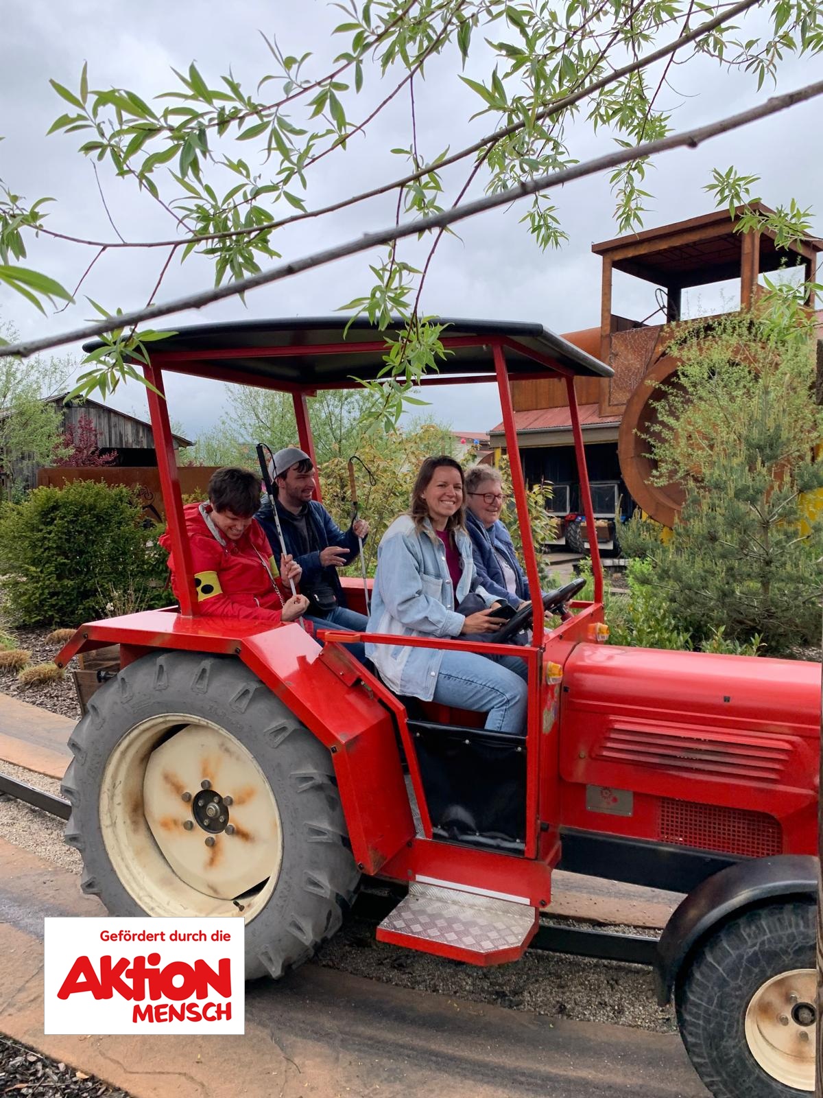 Roter Traktor mit überdachtem Anhänger auf einer Schiene in einer Parklandschaft.   Auf dem Fahrersitz vorne rechts sitzt eine junge Frau mit mittellangen hellbraunen Haaren, hellerer Haut, hellblauer Jeansjacke und blauer Jeans; sie lächelt deutlich.   Direkt hinter ihr sitzt eine weitere Person mit heller Haut, Brille, kurzem hellbraunen Haar und dunkelblauer Jacke, die nach vorne schaut.   Links davon sitzen zwei weitere Personen nebeneinander: vorn eine Person mit heller Haut, dunklen kurzen Haaren, roter Jacke und gelbem Armband am linken Oberarm, die nach unten zu einem Gegenstand in den Händen blickt; dahinter eine Person mit heller Haut, dunkler Kappe, dunkelblauer Jacke und Bart, die seitlich lächelt.  